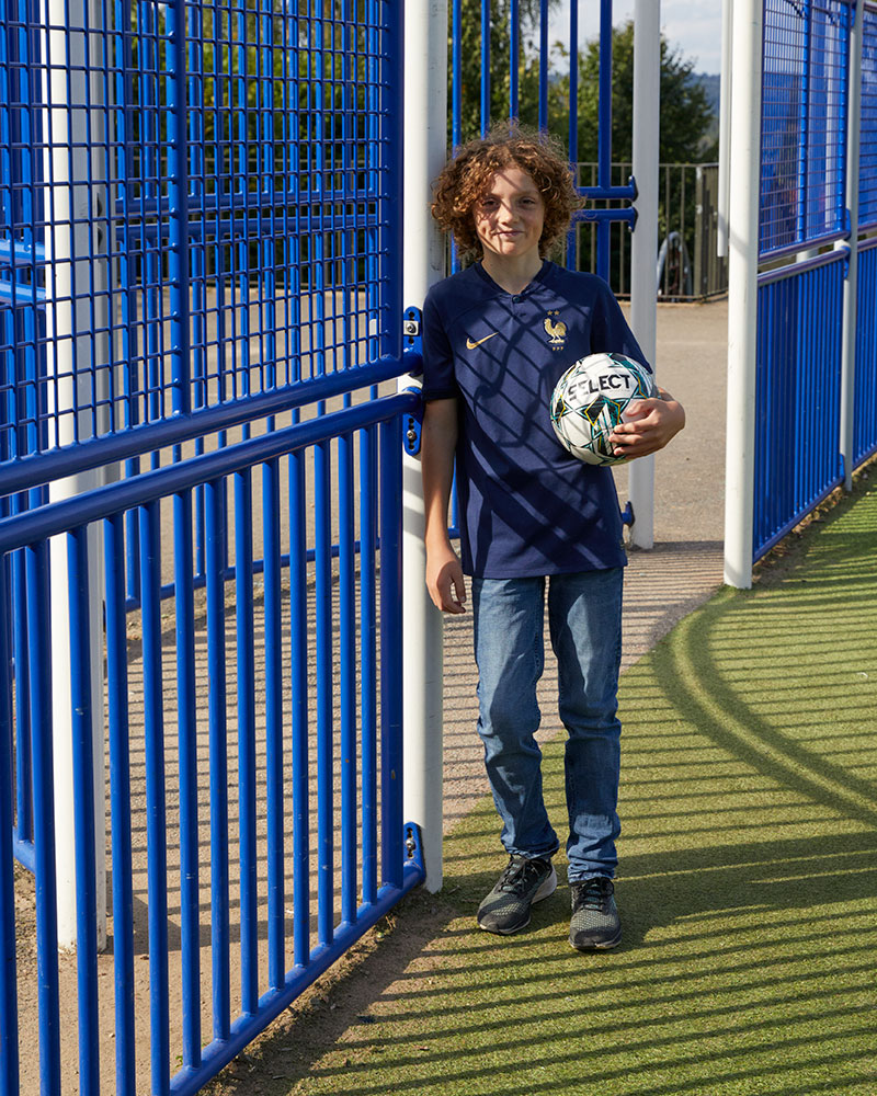 A boy poses with a football leaning against fence of a MUGA.